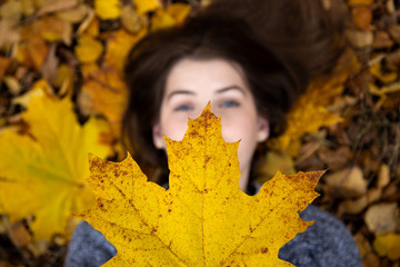 Top view of a cute girl with blue eyes, which in the fall lies on the ground and holds a beautiful yellow maple leaf in front of her. Face out of focus