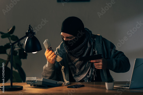 Fotografía Criminal in black mask holging pistol and looking at handset