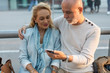 © Westend61 - Spain, Barcelona, senior couple sitting at tram stop in the city sharing smartphone with earbuds