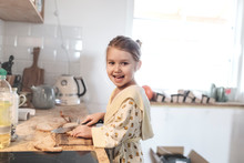 Portrait Of Laughing Little Girl Slicing Bread In The Kitchen