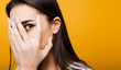 Close up portrait of a cute young european girl hiding face with her hand while looking at camera through fingers in front of a yellow studio background.