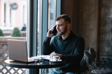 Young businessman talking on mobile phone while working on laptop in cafe