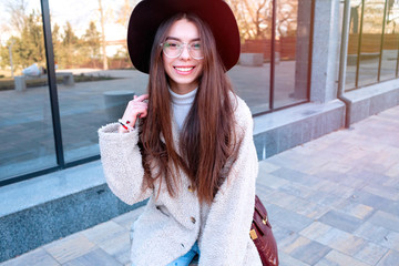 Outdoors lifestyle fashion portrait of happy stunning young girl. Beautiful smile. Walking to the city street. Long brunette hair. Wearing stylish beige coat. Joyful and cheerful woman. Happiness