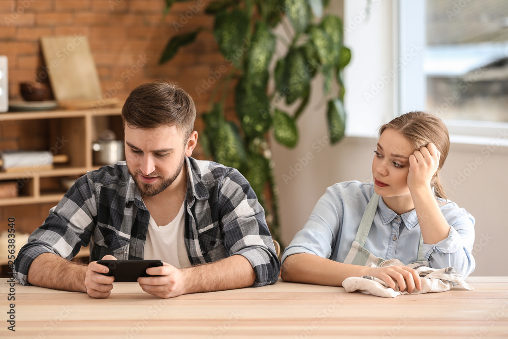 Lazy husband and sad wife sitting at table in kitchen