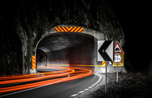 A Long Exposure Photo Showing The Entrance To The Tunnel In Mountain With Lots Of Light Tails And Several Traffic Signs Along The Road By Night.