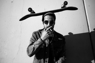 man posing on a urban wall smoking and wearing sunglasses with a skate on his head