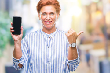 Sticker - Atrractive senior caucasian redhead woman showing smartphone screen over isolated background pointing and showing with thumb up to the side with happy face smiling