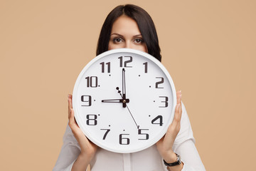 Photo of young modern business woman hold a clock in front of the face over beige background.