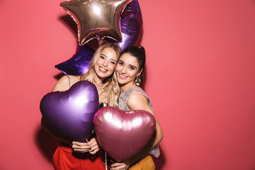 Image of two joyful girls 20s in stylish outfit laughing and holding festive balloons