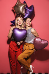 Image of two caucasian girls 20s in stylish outfit laughing and holding festive balloons