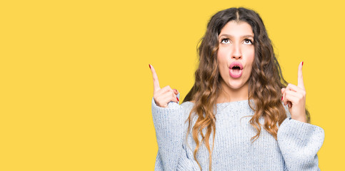 Young beautiful woman wearing winter sweater amazed and surprised looking up and pointing with fingers and raised arms.