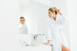 © LIGHTFIELD STUDIOS - selective focus of attractive and blonde woman in white bathrobe looking at mirror in bathroom