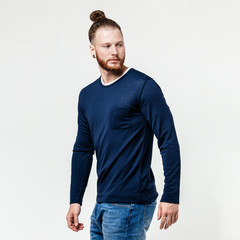 Wall Mural - Stylish man with beard and bun hairstyle dressed in blue long sleeve sweater and jeans poses in the studio on the white background