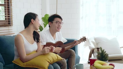 Wall Mural - Two of young Asian couple family relaxing at sofa playing guitar music and sing songs, Asian lifestyle at home concept