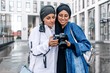 © pantovich - Two smiling girls in a hijab hugging watching photos on the camera standing on the street of the city