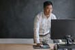 © mavoimages - Asian businessman standing at his desk working on a computer