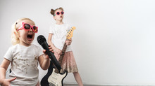 Two Girls In Pink Sunglasses And Identical T-shirts Sing With A Microphone And Play The Electric Guitar.