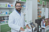 Fototapeta Sawanna - Cheerful mature male druggist smiling joyfully to the camera, standing behind checkout counter at his drugstore. Happy bearded chemist enjoying working at pharmacy. Medicine, sales concept