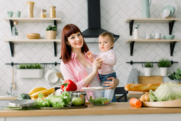 Funny picture of cute little girl with her mom at the table in the kitchen and looking at camera. Young beautiful woman with daughter cooking fresh salad at home.