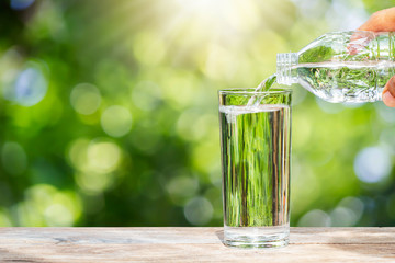 Wall Mural - Hand holding drinking water bottle pouring water into glass on wooden tabletop on blurred fresh green bokeh background