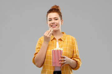 fast food and people concept - smiling red haired teenage girl in checkered shirt eating popcorn from striped bucket over grey background