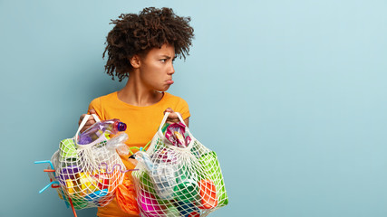 Dissatisfied offended dark skinned woman looks angrily aside, purses lower lip, holds two bags of plastic trash, has Afro haircut, stands over blue background with free space aside for advertisement