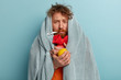 Photo of ill bearded man wrapped in coverlet, hold citrus and hot water bottle, measures temperature, trembles from cold, stay at home, has sick look, tries to break fever takes remedies. Seasonal flu