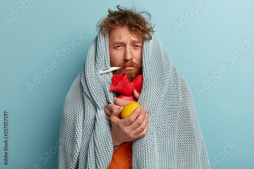 Fotografía Photo of ill bearded man wrapped in coverlet, hold citrus and hot water bottle, measures temperature, trembles from cold, stay at home, has sick look, tries to break fever takes remedies