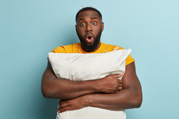 Morning and oversleep concept. Emotive stupefied dark skinned bearded guy embraces white soft pillow, gets up at night after seeing nightmare, stands over blue background, has feathers on body