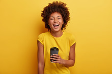 Monchrome Shot Of Cheerful Dark Skinned Woman Laughs And Talks Casually, Holds Disposable Cup Of Tea, Wears Yellow Bright T Shirt. Curly Afro American Girl Enjoys Hot Chocolate. Lifestyle Concept
