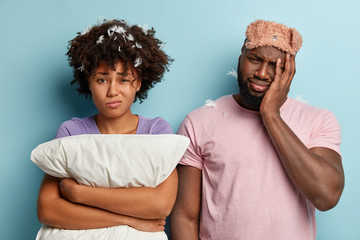 Tired ethnic man has problems with sleeping, cant get asleep, wears eyemask, frustrated dark skinned woman with pillow frowns face in dissatisfaction, isolated on blue background. Sleep and stress