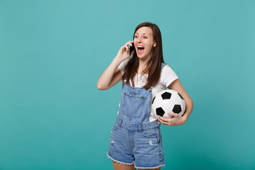 Excited cheerful young girl football fan with soccer ball talking on mobile phone conducting pleasant conversation isolated on blue turquoise background. People emotions, sport family leisure concept.