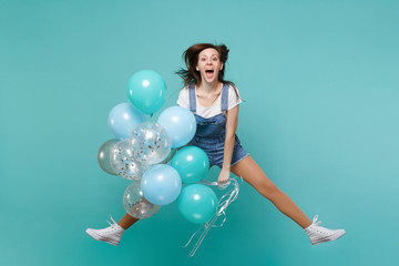 Cheerful young woman in denim clothes keeping mouth open, jumping high, celebrating, hold colorful air balloons isolated on blue turquoise background. Birthday holiday party, people emotions concept.