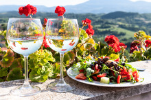 Closeup Of Arugula Salad With Olives And White Wine Two Glassses Plate On Balcony Terrace By Red Geranium Flowers In Garden Outside In Italy In Tuscany