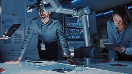 Poster - Diverse Team of Engineers with Laptop and a Tablet Analyse and Discuss How a Futuristic Robotic Arm Works and Moves a Metal Object. They are in a High Tech Research Laboratory with Modern Equipment.