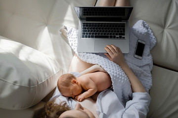 Cheerful young beautiful mother works as a freelancer at home, does remote work, breastfeeds her little child and reads information on a laptop.