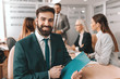 © Dusan Petkovic - Smiling bearded Caucasian businessman in formal wear holding pen and folder while sitting in boardroom. In background colleagues talking. Wish for it and work for it.
