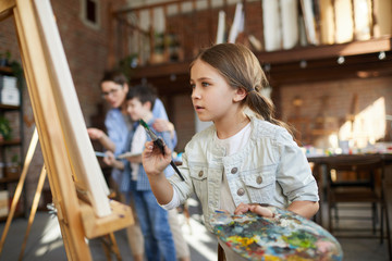 Wall Mural - Side view portrait of focused little girl painting picture on easel in art class and holding palette, copy space