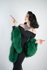 Wall Mural - charming graceful pin up girl in a black evening dress and a green fluffy boa in her hands stands and smokes a cigarette with a mouthpiece on a white solid Studio background