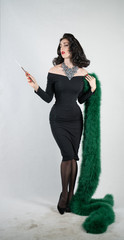 Wall Mural - charming graceful pin up girl in a black evening dress and a green fluffy boa in her hands stands and smokes a cigarette with a mouthpiece on a white solid Studio background