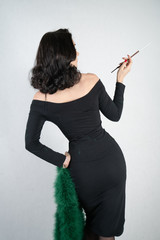 Wall Mural - charming graceful pin up girl in a black evening dress and a green fluffy boa in her hands stands and smokes a cigarette with a mouthpiece on a white solid Studio background