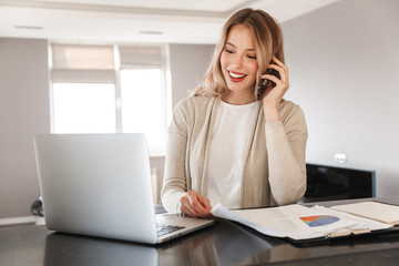 blonde woman posing sitting indoors at home using laptop computer talking by mobile phone.