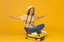 Young Dreamy Female Student Tourist Sitting On Suitcase, Pretending Flying Like Airplane, Isolated On Yellow Background. Dreams About Traveling Concept