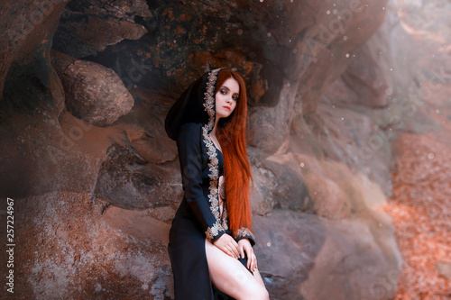 Cuadros en Lienzo mysterious young attractive lady in a long black cloak with a hood sits alone in