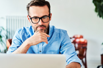 Thoughtful adult man with glasses using laptop at cafe