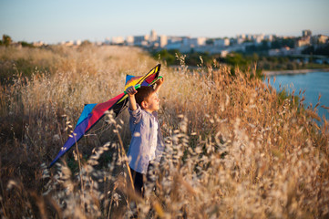 happy dreamy little caucasian boy with closed eyes holding kite standing among field near sea and city