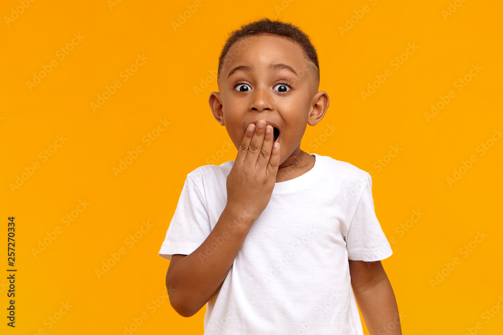 Wow. Studio shot of emotional adorable African American little boy raising eyebrows and covering open mouth with hand being surprised and shocked, showing true astonished reaction on unexpected news - obrazy, fototapety, plakaty 