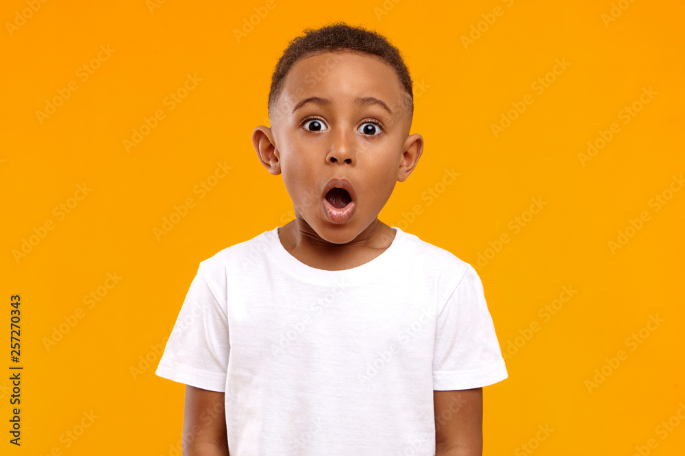Surprise, excitement and fascination concept. Funny bug eyed African little boy opening his mouth widely, shocked with astonishing unexpected news, having amazed look, showing full disbelief - obrazy, fototapety, plakaty 