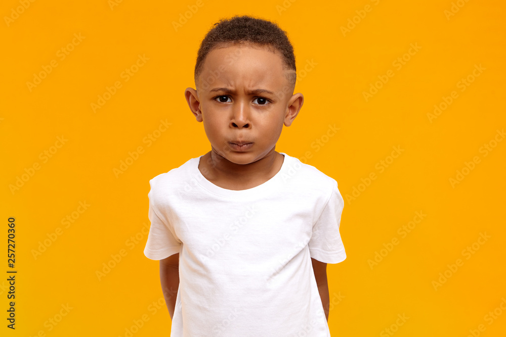 Studio image of displeased frowning Afro American little boy in white t-shirt having grumpy dissatisfied facial expression, being grounded by parents for bad behavior. Human emotions and reaction - obrazy, fototapety, plakaty 