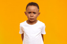 Studio Image Of Displeased Frowning Afro American Little Boy In White T-shirt Having Grumpy Dissatisfied Facial Expression, Being Grounded By Parents For Bad Behavior. Human Emotions And Reaction